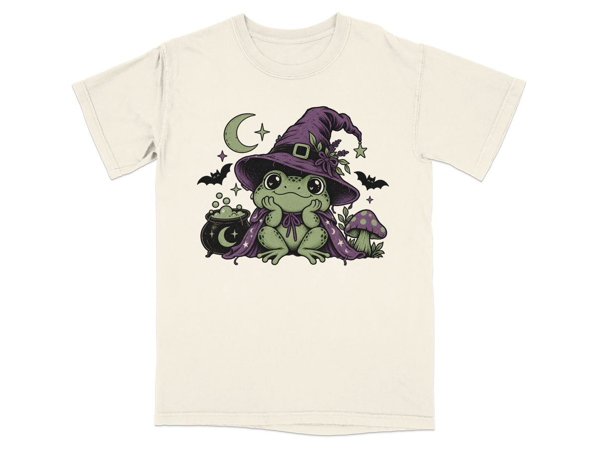 A cream-colored t-shirt featuring an illustration of a cute green frog wearing a witch hat and cape.