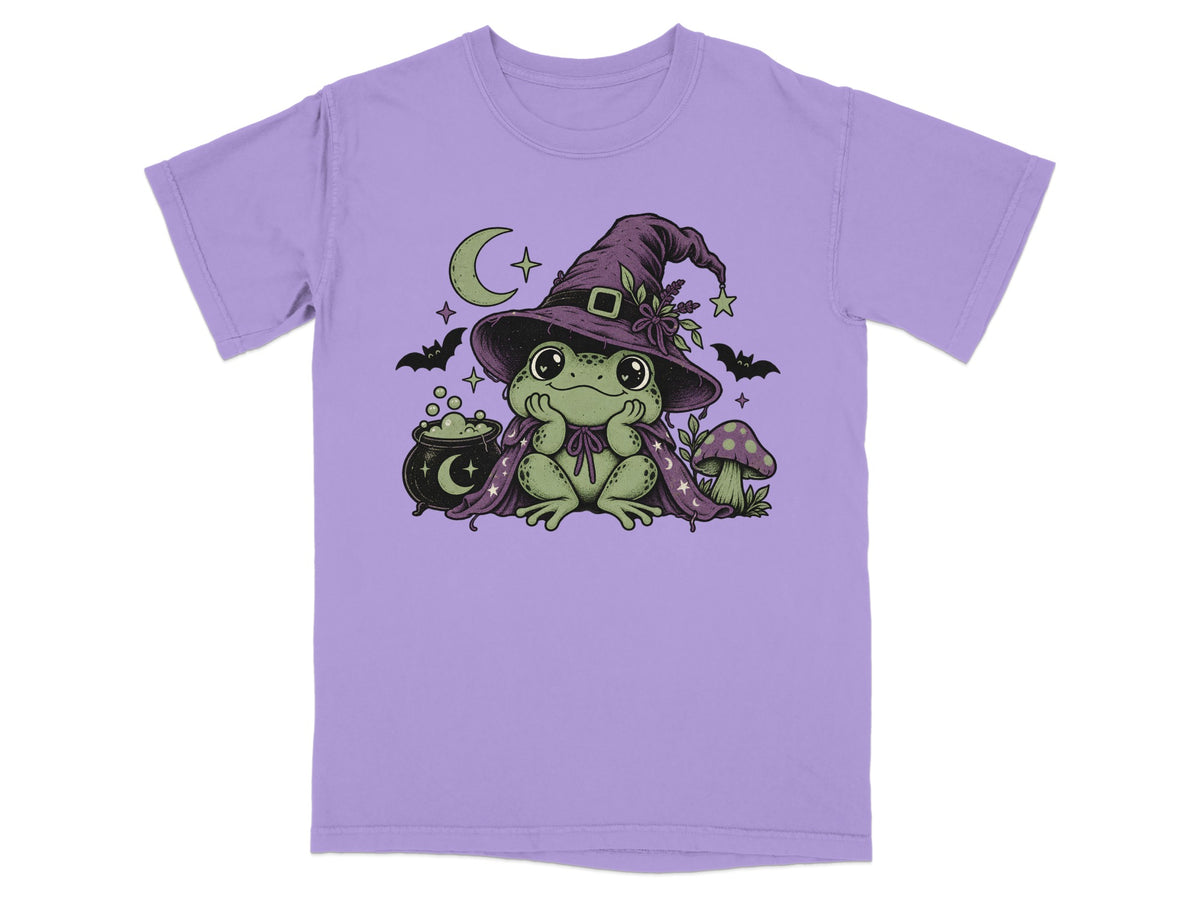 A purple t-shirt printed with a cute frog wearing a witch hat and cape, flanked by bats, a cauldron, and a mushroom.