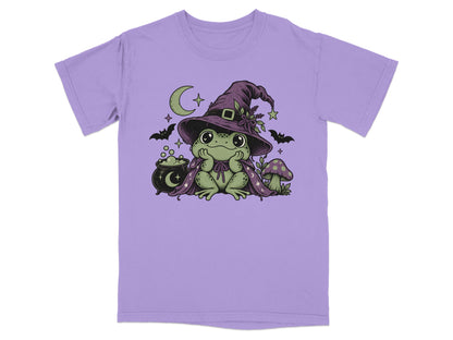 A purple t-shirt printed with a cute frog wearing a witch hat and cape, flanked by bats, a cauldron, and a mushroom.