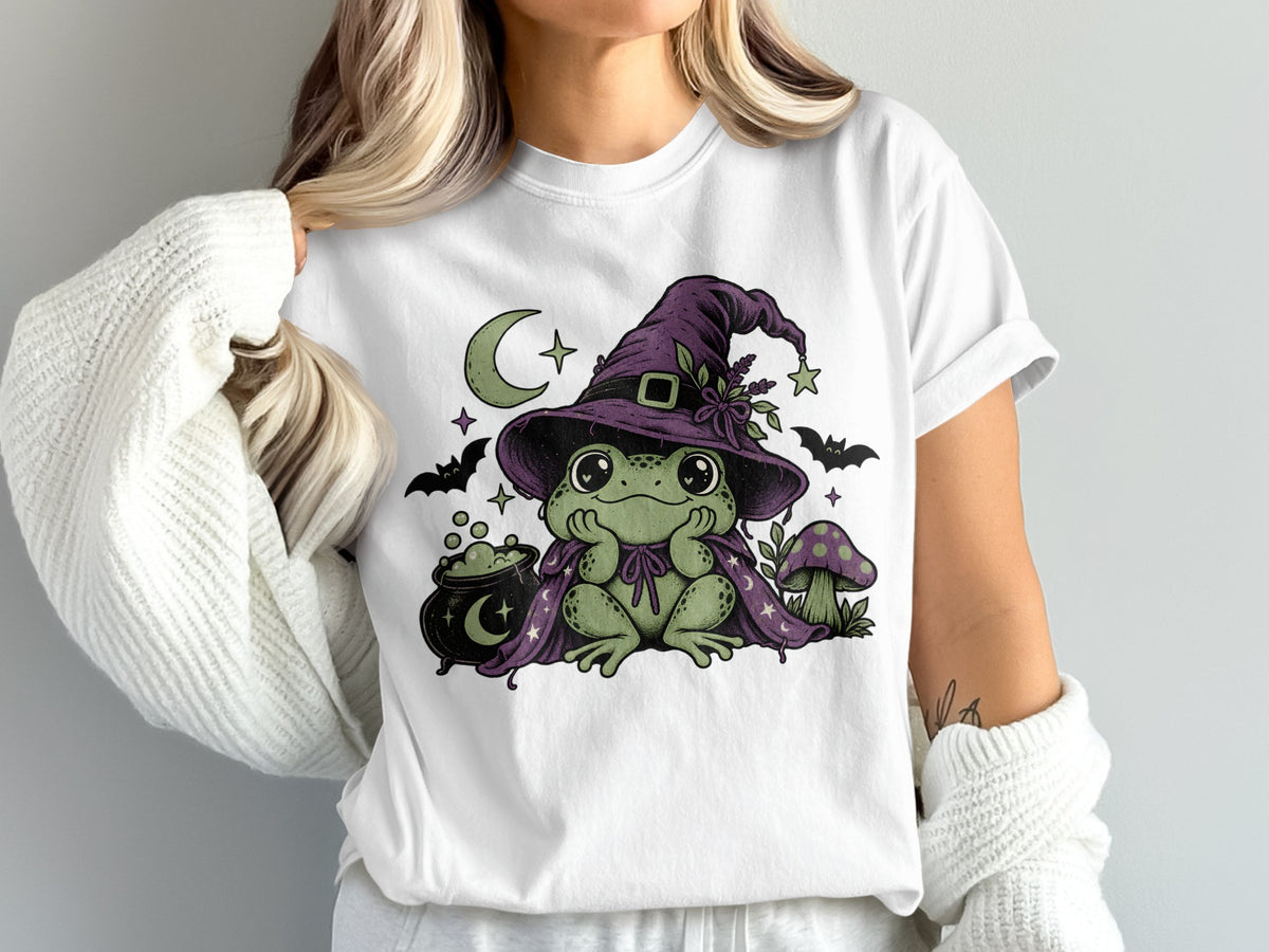 A person wears a white t-shirt featuring a green frog wearing a purple witch hat and cape, surrounded by cauldrons, bats, and mushrooms.