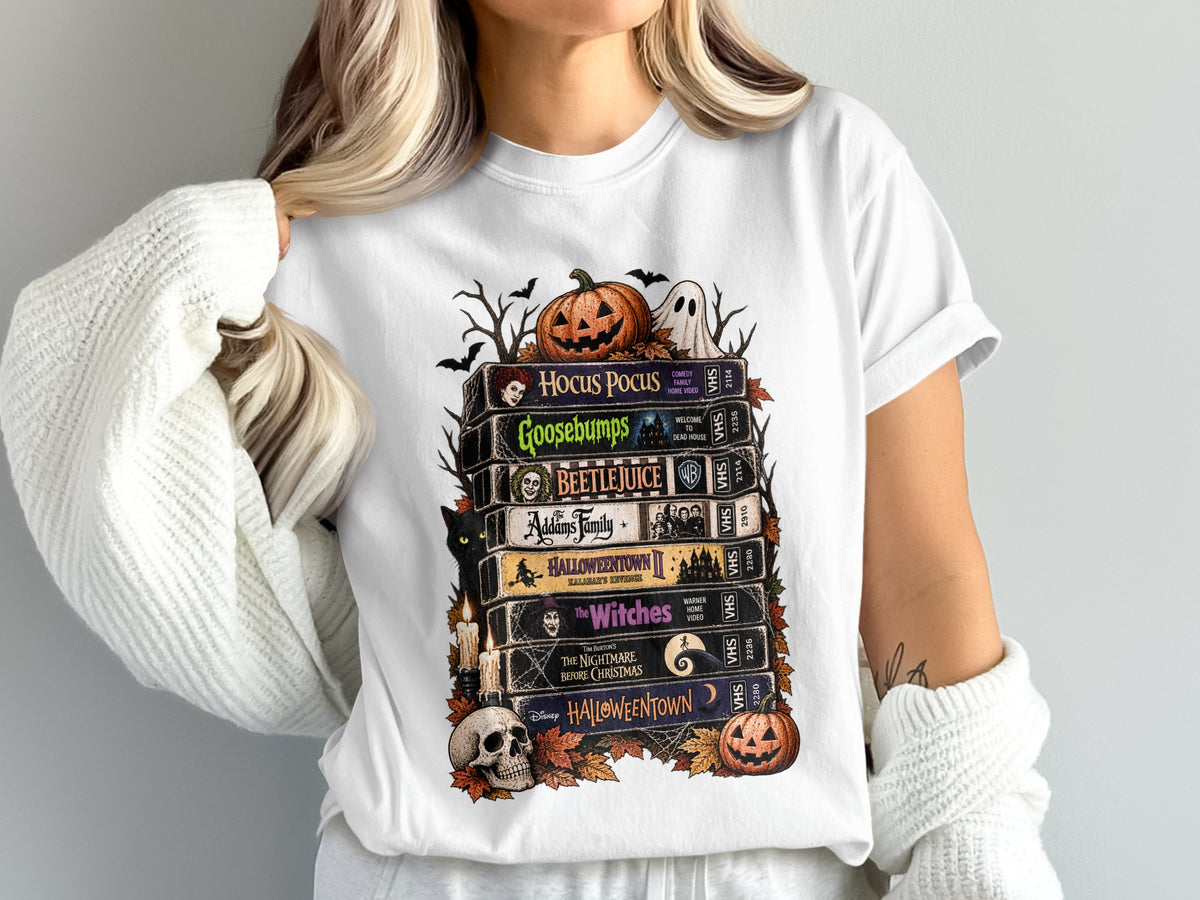 White t-shirt featuring stack of Halloween VHS tapes: Hocus Pocus, Goosebumps, Beetlejuice, Addams Family, Halloweentown II, The Witches, Nightmare Before Christmas.