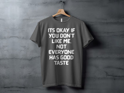 Dark grey t-shirt with white text reading "IT'S OKAY IF YOU DON'T LIKE ME. NOT EVERYONE HAS GOOD TASTE."