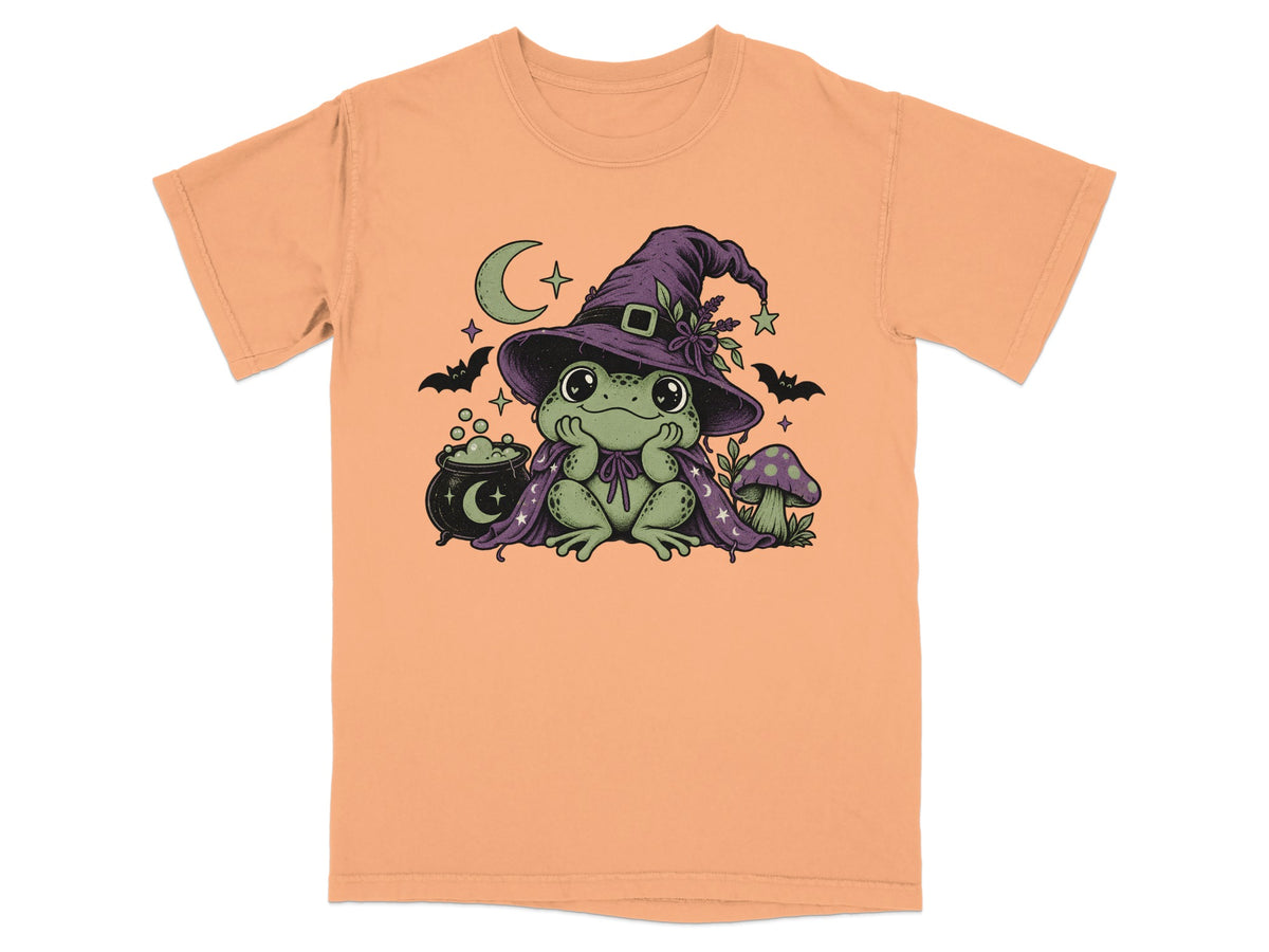 Peach-colored t-shirt featuring a green frog wearing a witch hat and cape, with a cauldron and mushroom.