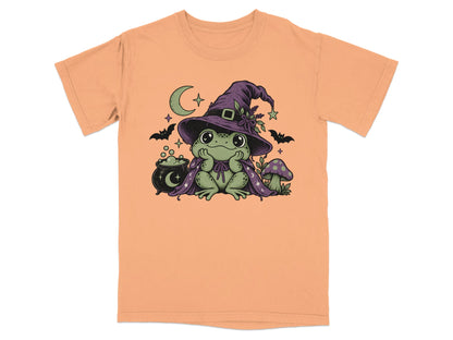Peach-colored t-shirt featuring a green frog wearing a witch hat and cape, with a cauldron and mushroom.