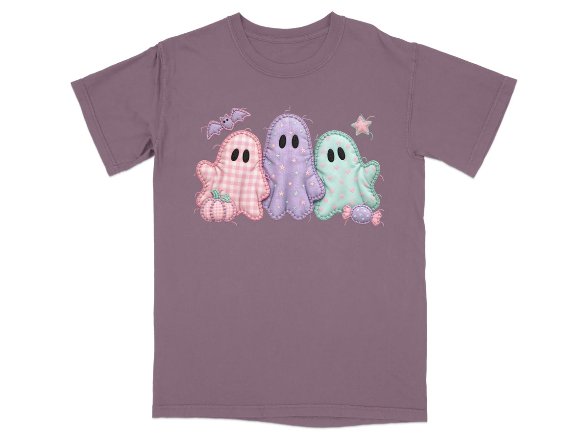 Three pastel Halloween ghosts with a bat, pumpkin, and candy decorations on a purple t-shirt.