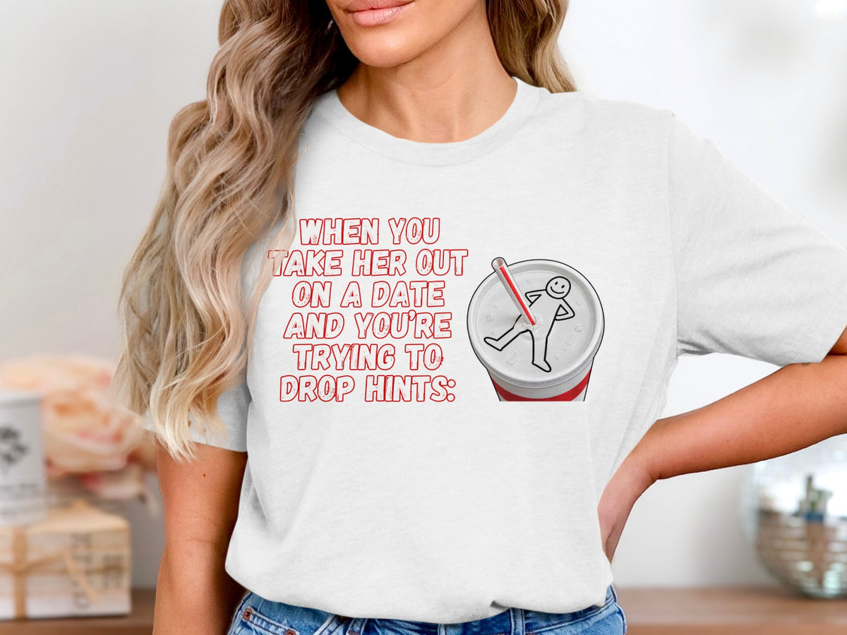 White t-shirt with red text "WHEN YOU TAKE HER OUT ON A DATE AND YOU'RE TRYING TO DROP HINTS:" and graphic.