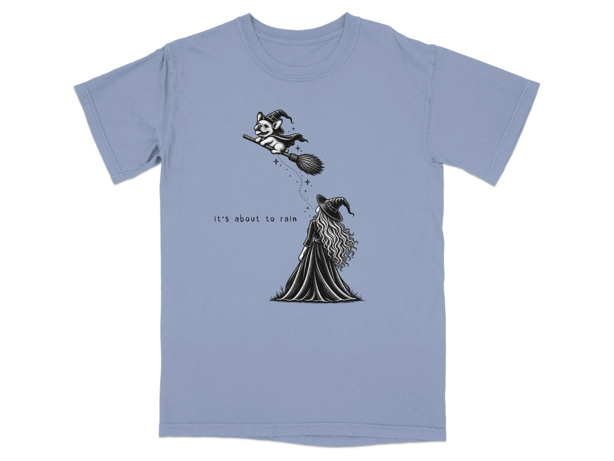 A slate blue t-shirt printed with a black and white illustration of a witch looking up at a dog flying on a broomstick, with the text It's about to rain.
