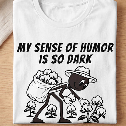White t-shirt with black text "MY SENSE OF HUMOR IS SO DARK" and cartoon figure picking cotton.