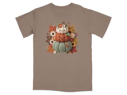 Three stacked knitted pumpkins with smiling faces surrounded by autumn leaves and flowers.