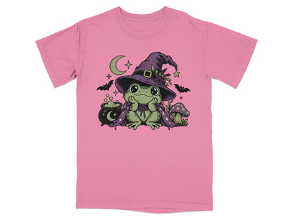 A pink t-shirt featuring a green frog wearing a witch hat and cape, surrounded by a crescent moon, bats, a cauldron, and mushrooms.