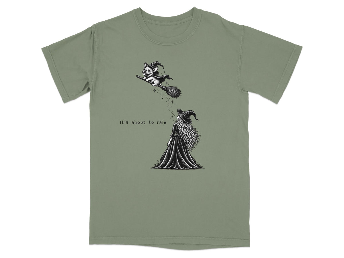 An olive green t-shirt featuring a black and white illustration of a witch looking up at a dog flying on a broomstick, with the text "It's about to rain".