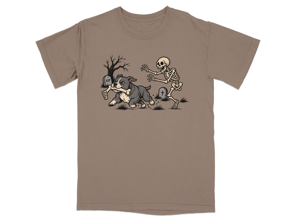 A tan t-shirt printed with a cartoon graphic of a skeleton chasing a dog running away with a bone.
