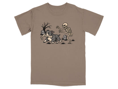 A tan t-shirt printed with a cartoon graphic of a skeleton chasing a dog running away with a bone.