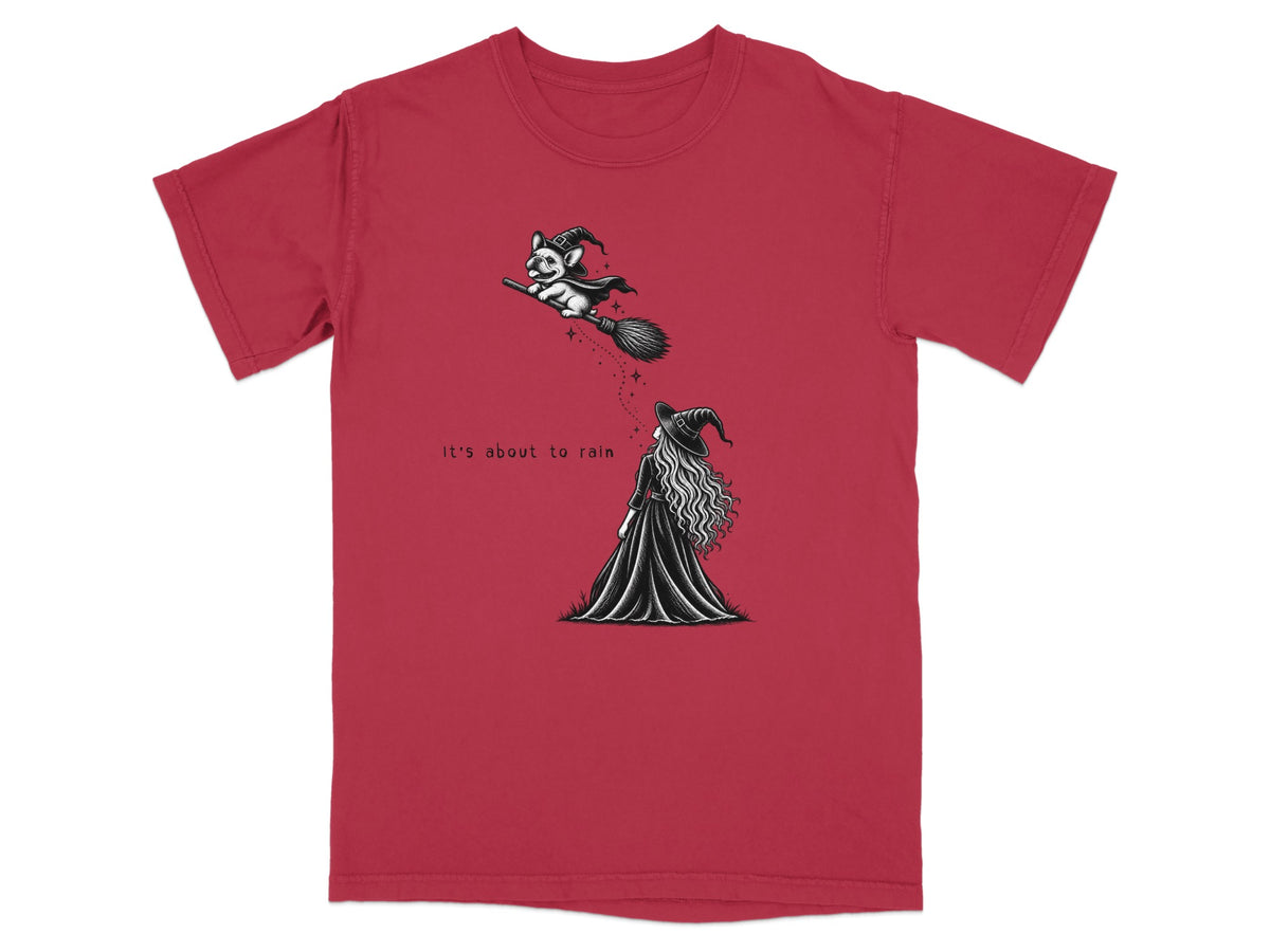 A red t-shirt with a black and white graphic of a witch watching a dog on a broomstick, featuring the text "It's about to rain".