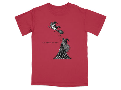 A red t-shirt with a black and white graphic of a witch watching a dog on a broomstick, featuring the text "It's about to rain".
