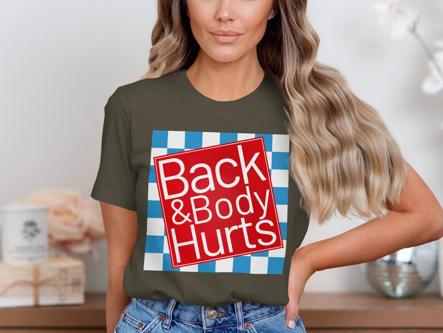Olive green t-shirt with red square graphic reading "Back & Body Hurts" on checkered background.