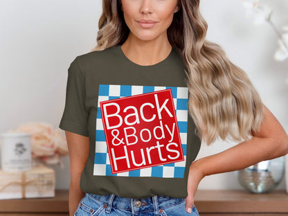 Olive green t-shirt with red square graphic reading "Back & Body Hurts" on checkered background.