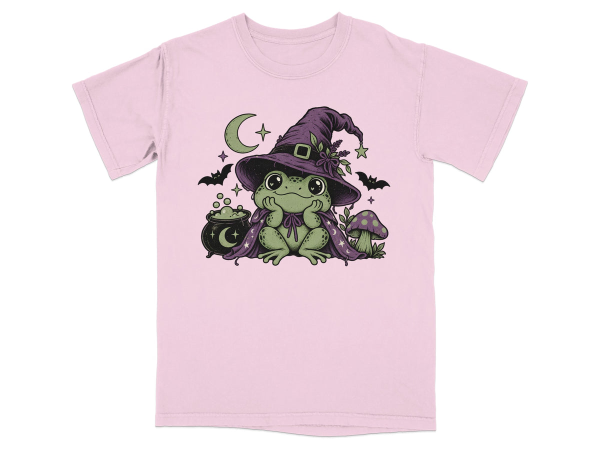 Light pink t-shirt featuring a green frog wearing a witch hat and cape, with a cauldron and mushroom.
