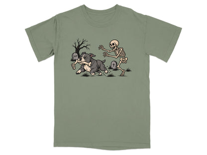 An olive green t-shirt featuring a cartoon design of a dog running away from a chasing skeleton in a graveyard.