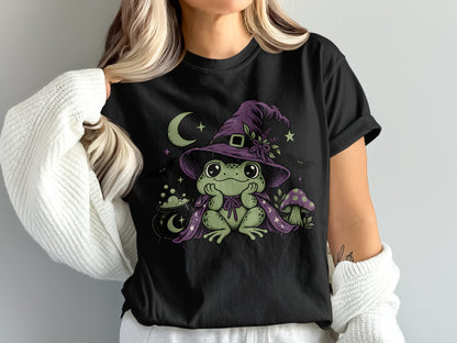 A black t-shirt featuring a green frog wearing a purple witch hat and cape, with a cauldron and mushroom.