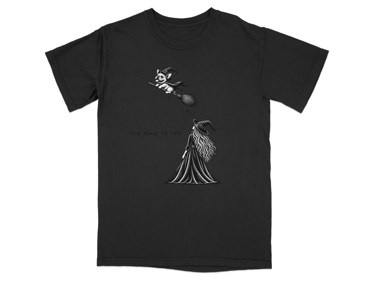 A black short-sleeved t-shirt featuring a white illustration of a witch looking up at a dog flying on a broomstick, with the text It's about to rain.