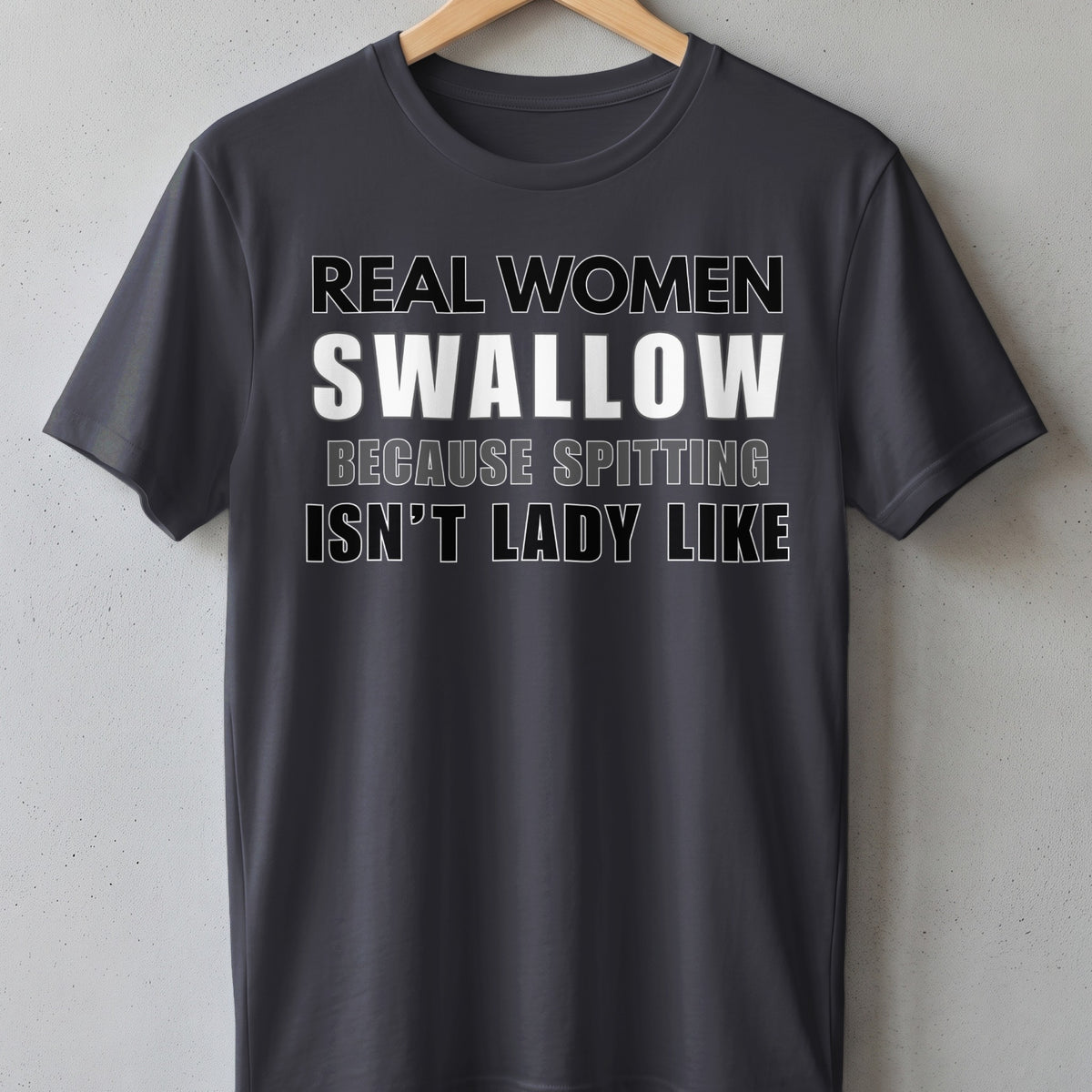 Dark grey t-shirt with white and grey text reading "REAL WOMEN SWALLOW BECAUSE SPITTING ISN'T LADY LIKE".