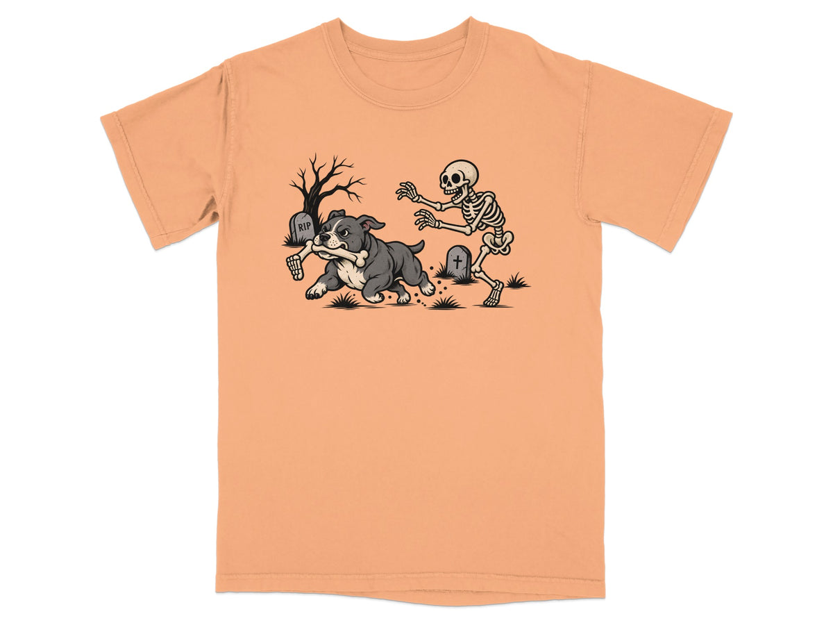 Peach-colored t-shirt featuring a graphic of a bulldog running away from a skeleton in a graveyard.