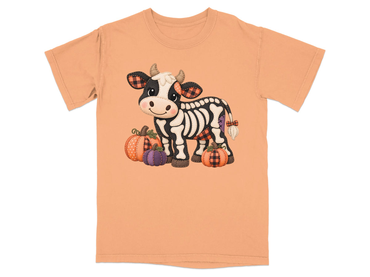 Skeleton cow with pumpkins and plaid accents on a peach t-shirt.