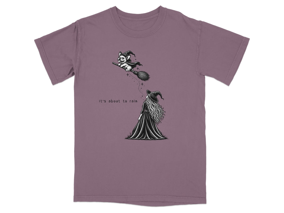 A muted purple crewneck t-shirt featuring a black and white graphic of a witch and a dog riding a broomstick with the text "it's about to rain".