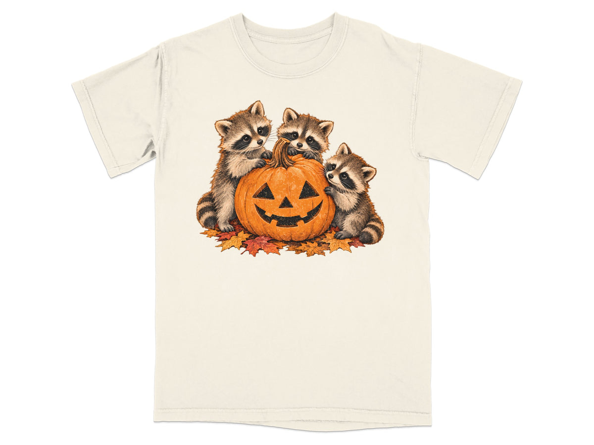 Three raccoons with a jack-o'-lantern and autumn leaves on a cream t-shirt.