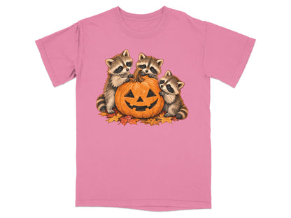 Three raccoons surround a jack-o'-lantern on a pink t-shirt.