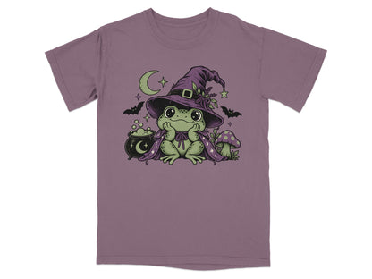 A mauve t-shirt featuring a green frog wearing a witch hat and cape, surrounded by a cauldron, bats, and a crescent moon.