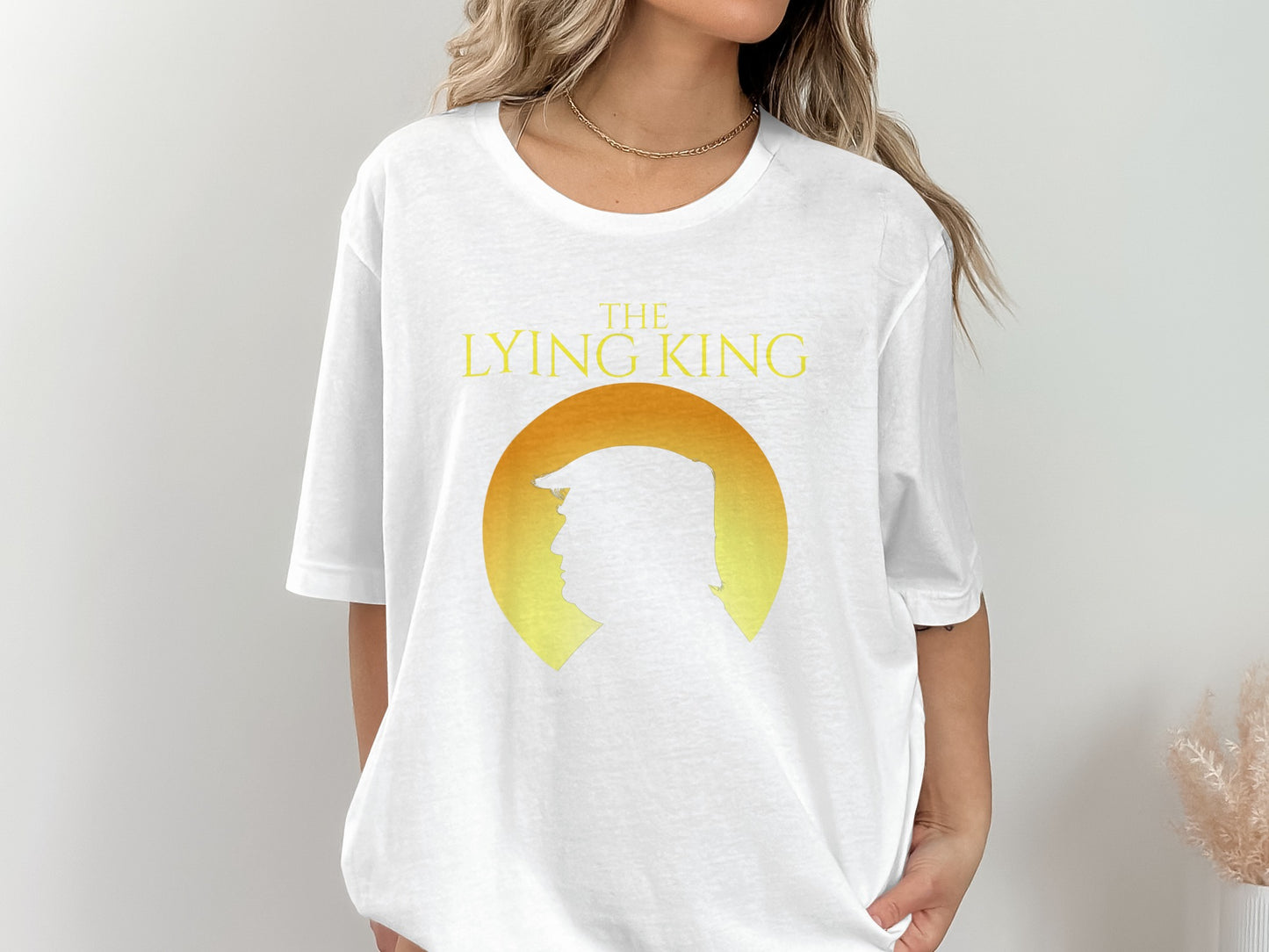 White t-shirt with yellow text "THE LYING KING" and silhouette of Donald Trump.