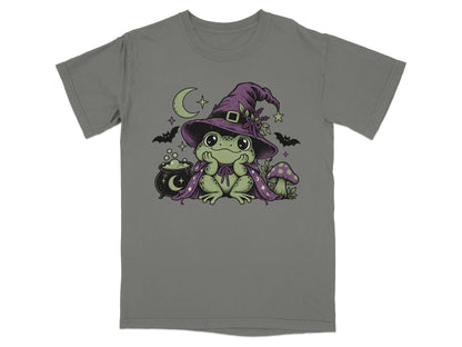 A grey short-sleeved t-shirt featuring a green frog wearing a purple witch hat and cape, surrounded by a crescent moon, bubbling cauldron, mushrooms, and bats.