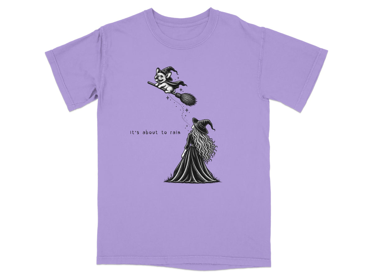 Light purple t-shirt featuring a black graphic of a witch and a flying dog with text reading, "It's about to rain."
