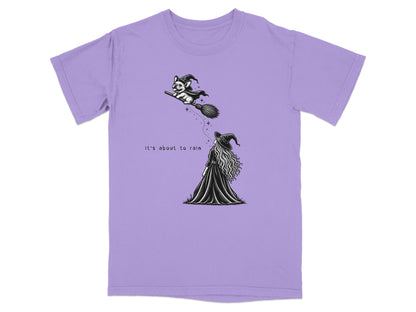 Light purple t-shirt featuring a black graphic of a witch and a flying dog with text reading, "It's about to rain."