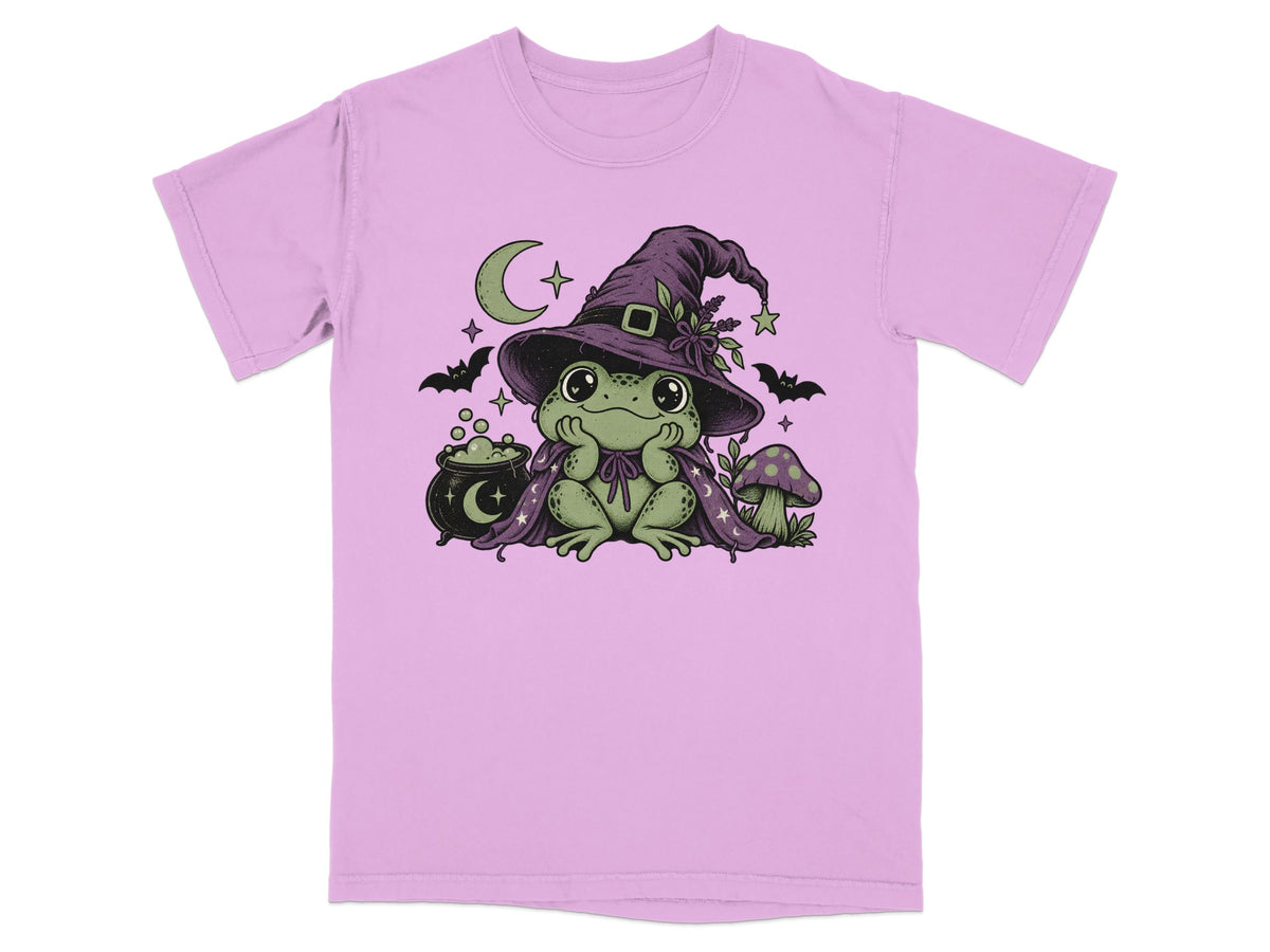 A light purple t-shirt featuring a green frog wearing a witch hat and cape, surrounded by cauldrons, bats, and stars.