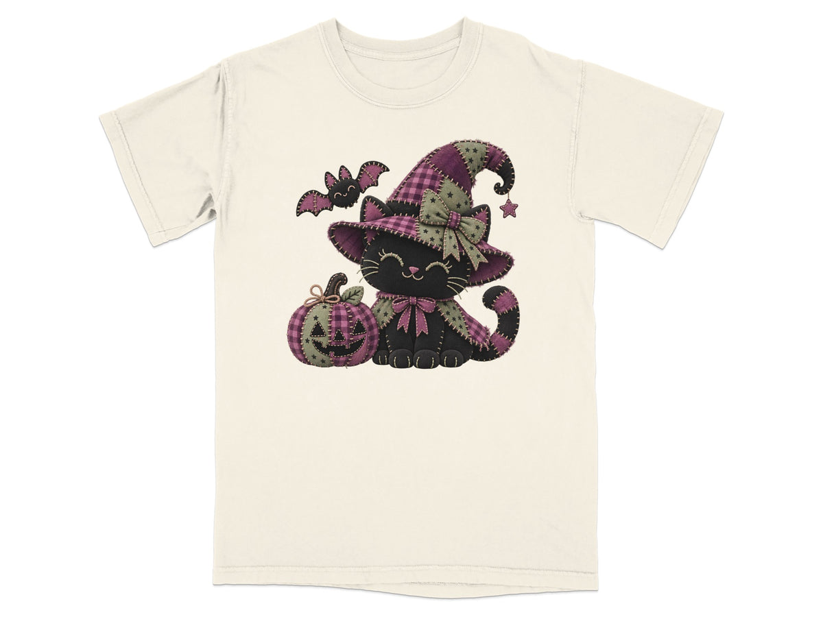 An off-white short-sleeve t-shirt with a graphic of a black cat wearing a purple witch hat, sitting next to a jack-o-lantern.
