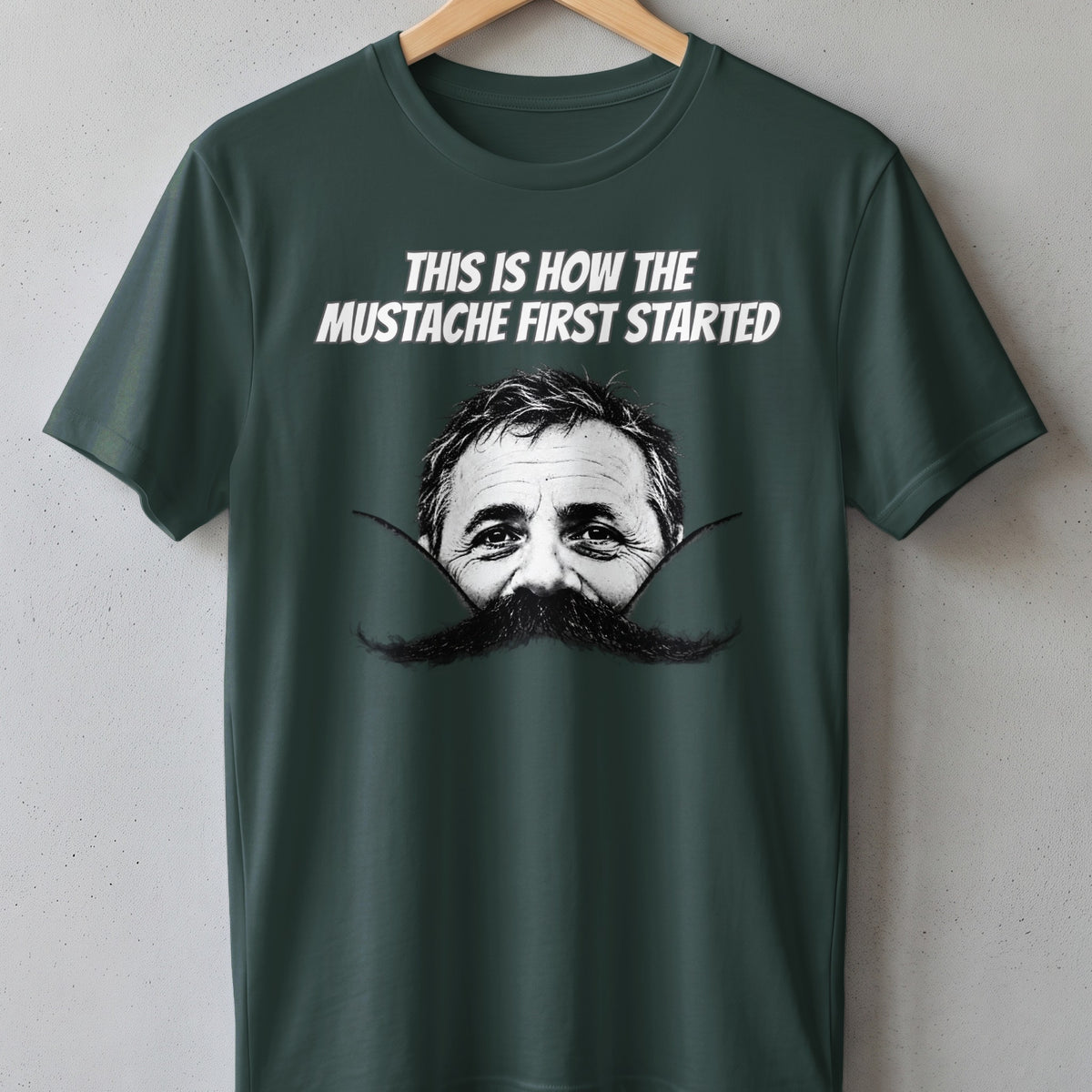 Dark green t-shirt with man's face and large mustache graphic, text reads "This is how the mustache first started."