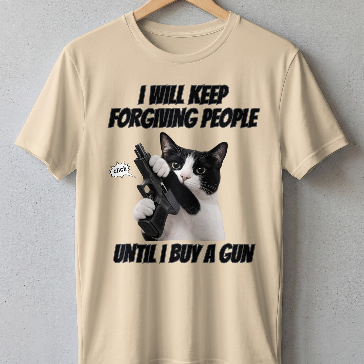 Cream t-shirt with a black and white cat holding a gun and text "I WILL KEEP FORGIVING PEOPLE UNTIL I BUY A GUN".