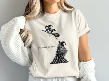A cream-colored t-shirt featuring a black line drawing of a witch and a dog on a broomstick, with the text "It's about to rain."