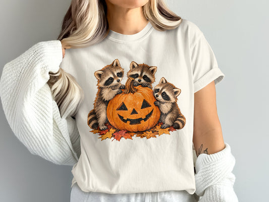 Three raccoons sit with a jack-o'-lantern surrounded by autumn leaves.