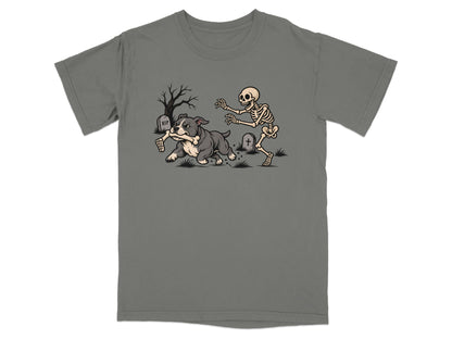 A grey t-shirt featuring a graphic of a skeleton chasing a bulldog holding a bone in a graveyard.
