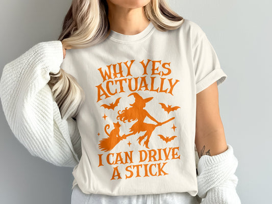 Cream t-shirt with orange graphic of witch, cat, and bats, reading "Why Yes Actually I Can Drive A Stick".